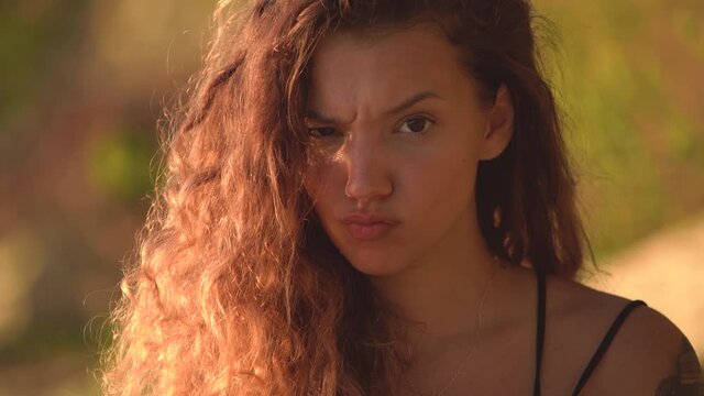 Young woman with curly hair raising eyebrow and making cute mad face, pouting expression. Close up shot and blurred nature background.