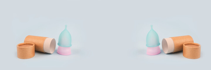 Blue and pink menstrual cups on light background, with paper packaging tube - copyspace and mockup. Women health and zero waste concept. Female hygiene period products, alternative lifestyle. Banner
