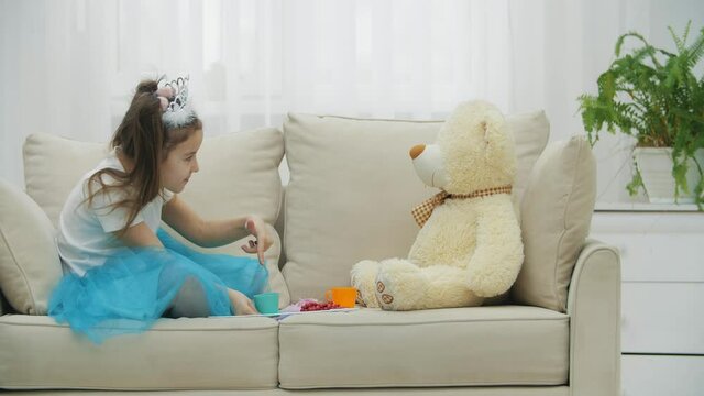 Cute girl in crown and dress, teaching her best friend Teddy Bear how to drink tea. But toy is not alive and can't do it.
