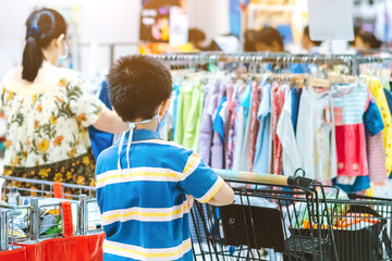 Back view of boy wearing face mask to prevent the spread of the Corona Virus (Covid-19) shopping in a supermarket. Boy with shopping cart during virus outbreak in grocery store. New normal lifestyle.