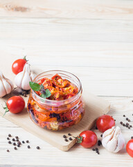 Sun dried tomatoes in glass jar with ripe tomatoes and garlic on cutting board, on wooden background