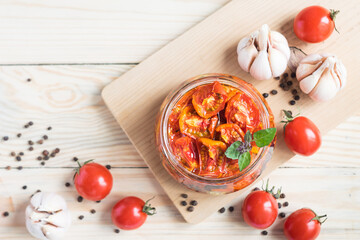 Sun dried tomatoes in glass jar with ripe tomatoes and garlic on cutting board, on wooden background, top view