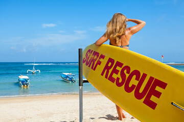 Life saving yellow board with surf rescue sign. Young lifeguard woman stand on duty, look at blue...