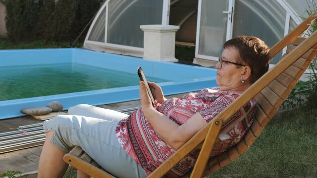 An elderly woman is sitting on a wooden deck chair by the pool in the backyard and enjoys reading an e-book.