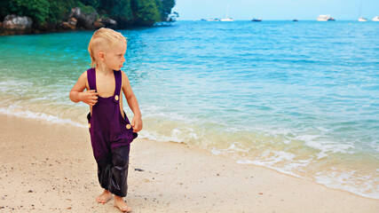 Happy barefoot child in loose jumpsuit have fun on tropical sea beach. Funny baby boy run by water along surf edge. Active kids lifestyle, people activity on family summer holiday with children