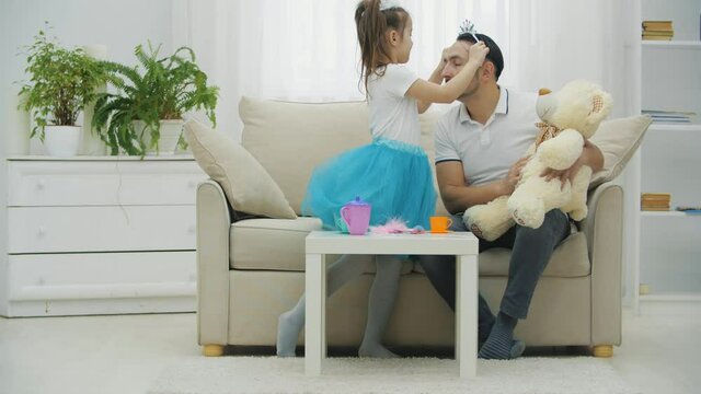 Cute princess girl making her father beautiful, putting crown on his head. Father is holding bear on his knees.