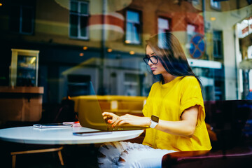 Fototapeta premium Thoughtful attractive female freelancer in cool spectacles keyboarding message for customer while making remote job indoors.Cute brunette blogger typing publication for personal website on laptop
