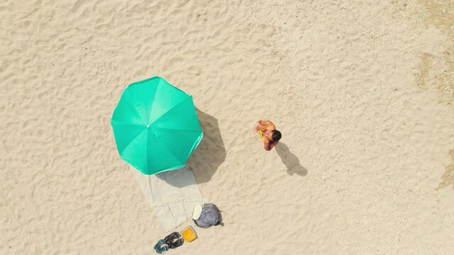 aerial drone zooming out footage beautiful sexy taned latin woman in bikini standing on sandy beach and combing her wet hair. clothes, backpack, beach mat and turquoise sun umbrella on sand