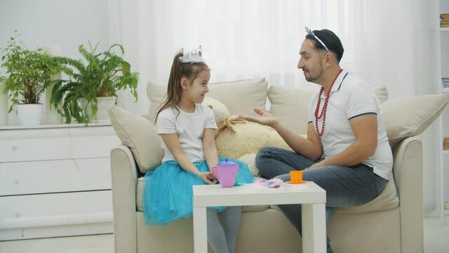 Father and daughter playing in princesses. Funny man kissing girl's hand, then pretending to bite it. He hugs her and breaks a crown. Tears on baby's eyes.