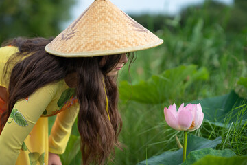 Beautiful girl with Vietnam culture traditional hat and dress, Ao dai, bent over a blossoming lotus flower. Portrait of Asian girl with Ao-Dai Vietnam traditional dress costume for woman. 