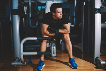 Fototapeta na wymiar Full length portrait of young athlete seated on gym equipment and taking a break after workout