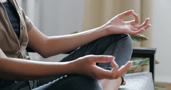 Woman wearing casual jeans clothes sitting on sofa in room in lotus position with crossed legs, doing yoga home, practicing asanas, gaining energy, relaxing, concentrate zen harmony concept, close up