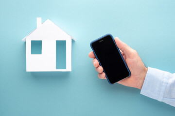 Model of a house made of paper, hand with mobile phone on a blue background, home work concept.