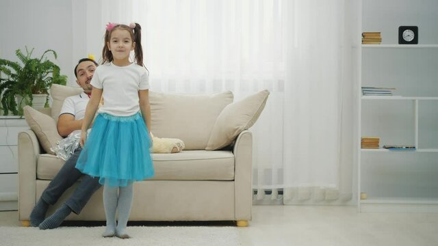 Little girl is turning around like a ballerine. Father is sitting on the sofa, looking at her, smiling.