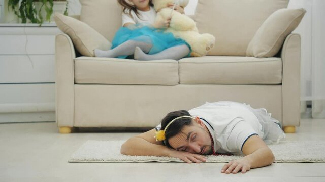Father in crown is lying on the floor completely exhausted and wants to sleep. His daughter is full of energy, sitting on the sofa with magic wand and teddy, playing.