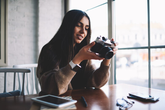 Attractive female with brown hair holding vintage camera in hand and watching funny images.Creative photographer enjoying taken pictures sitting in coffee shop interior  during leisure time