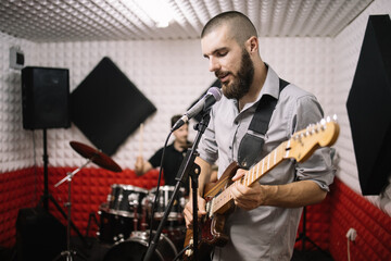Fototapeta premium Guitar player singing while playing in music studio. Rock band of guitarist and drummer rehearsing in recording room.