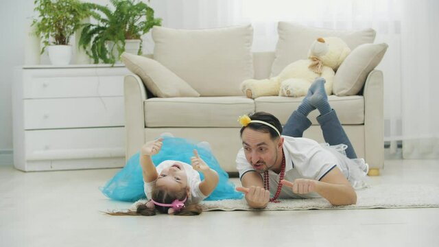 Cute father and daughter lying on the floor in princess clothes, father on the belly, daughter on the back, smiling, giving thumbs up.