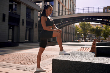 Beautiful slim stylish girl athlete, early morning workout does stretching muscles before jogging, free space text motivation fitness workout. Sportswear leggings top, sky background stairs buildings.