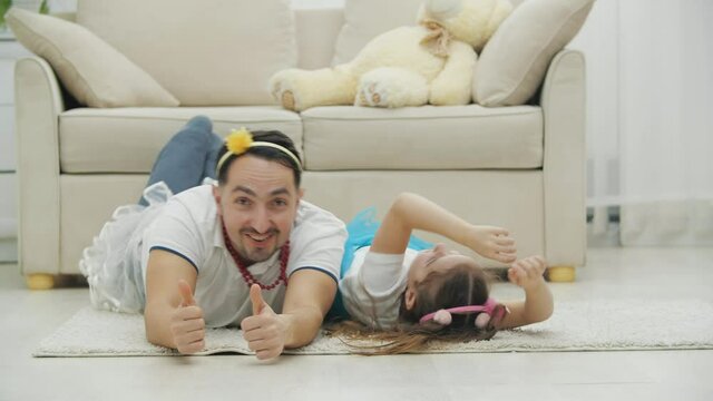 Lively father and daughter in funny headgears and skirts are playing, rolling on the floor, giving thumbs up, laughing.