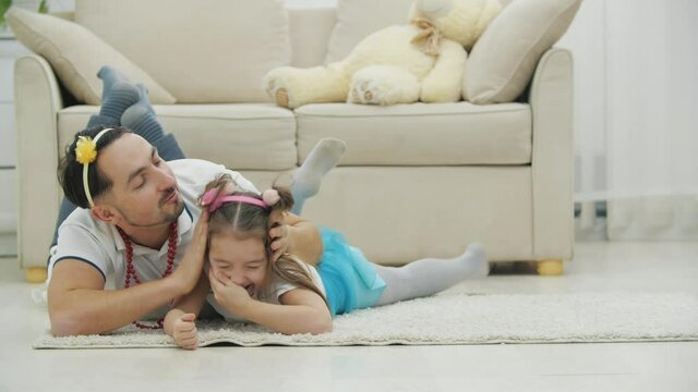 Affectionate father is kissing her little daughter on cheek while they are playing, lying on the floor, rolling, laughing.