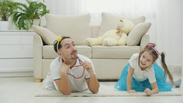 Funny father is posing lying on the floor, showing necklace, then giving five her little beautiful daughter that is rolling on the floor near him.