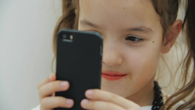 Cropped video of cute little girl with pigtails and necklace, holding black phone, taking photos.