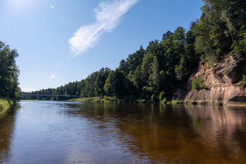 On a beautiful summer day, the river Gauja flows calmly on which floor the rock is reflected in the river water, the sky is blue with some clouds