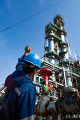 Oil refinery worker with one hand up. Blue work wear and helmet and grey oil distillation tower...