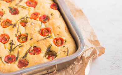 Traditional italian focaccia bread with tomatoes and rosemary in mental baking tray . On the beige brown paper and white concrete background.