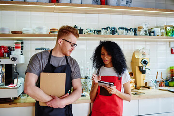Two diverse colleagues baristas dressed in aprons discussing order for own cafeteria standing against bar.African american young woman talking with caucasian waiter about menu during work in cafe