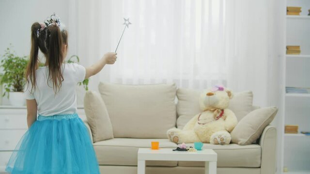 Little fairy standing with magic wand in her hand, conjuring her teddy bear on the sofa, then turning to the camera, waving wand in front of it, smiling.