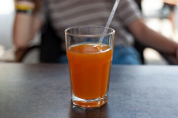 There is a glass of orange juice on the table in the cafe. There is a straw in the glass.