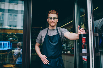 Portrait of successful caucasian barista in apron smiling at camera and inviting guests in coffee shop.Cheerful male waiter smiling at camera while standing at entrance to own cafeteria
