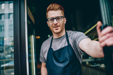 Portrait of positive caucasian male barista in eyeglasses smiling at camera while opening door in...