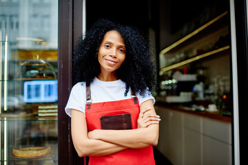 Half length portrait of cheerful african american professional barista in red apron smiling at camera.Positive dark skinned female waitress with crossed hands standing at entrance to own coffee shop