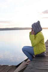 Model sitting on a boardwalk near a lake wearing yellow jacket with gray hood.