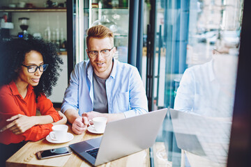 Two male and female business partners have meeting in coffee shop and talking about online database on modern laptop device, multicultural colleagues discussing collaborative plan for cafeteria