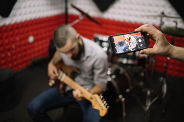 Male hand taking photo of guitarist using phone. Cropped human photographing man playing guitar and sitting in music recording studio.