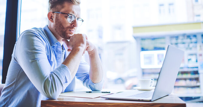 Serious male software developer working remotely in cafe interior with good wifi connection checking program code in laptop application and feeling pondering on mistakes, concept of distance job
