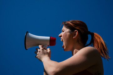 Red-haired Caucasian woman yelling into a megaphone against a blue sky background. The girl makes an announcement through the loudspeaker.