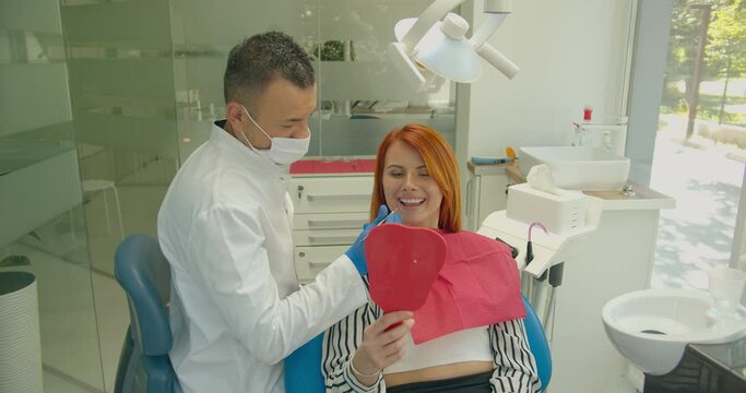 Professional male dentist show beautiful teeth in mirror to happy woman visitor. Positive red-haired female patient of dental clinic enjoying amazing smile after treatment.