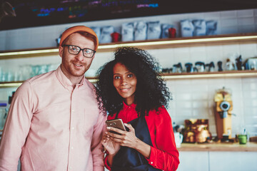 Portrait of cheerful caucasian owner of cafe standing together with african american female barista and smiling at camera.Two professional colleagues working in team in common coffee shop