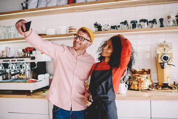 Successful male blogger making selfie photo with african american barista in black apron during working process in cafeteria.Positive caucasian tourist with waitress talking picture on front camera
