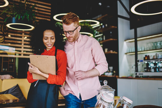 Positive african american waitress taking order in caucasian hipster guy during working process in coffee shop.Positive owner of cafe together with dark skinned barista discussing products