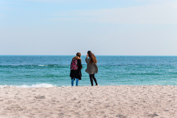 Young girlfriends stand with their backs on the beach and look at the sea
