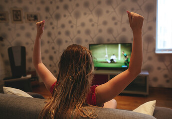 Image of a woman watching a football match on TVat home with raised hands.