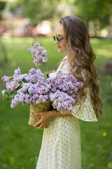 Fototapeta premium The girl in white dress and sunglasses holds in her hands a wicker basket with flowers. Basket with lilacs. Girl and flowers. Walk with a basket of lilacs in the hands. Floristics