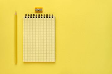 Yellow spring notepad and yellow pencil are on the left side of the screen on yellow background. Squared paper texture. Monochrome horizontal photo, flat lay, copy space.
