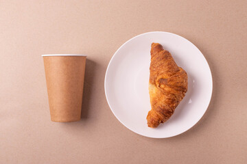a cardboard coffee cup and a croissant on beige rustic background